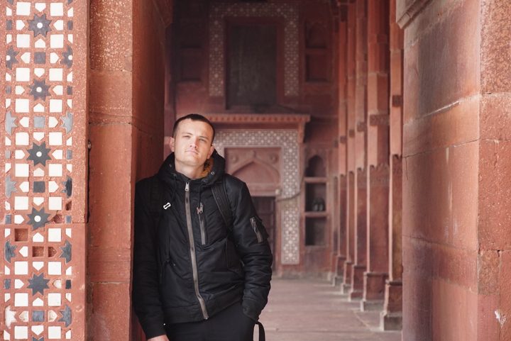 A person in a dark jacket stands among red stone arches and geometric patterned panels.